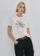 Remera de algodón color crudo, con cuello redondo, manga corta y estampa frontal de flores en tonos lilas con la leyenda "Self made ready to flourish".
