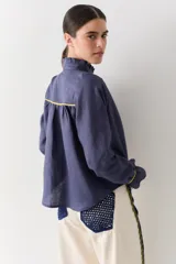 Camisa blanca de lino con cuello alto tipo gorguera, cierre frontal con botones de madera y detalles de ribetes en color mostaza en hombros y puños. Presenta volados en la botonera, cuello y puños.