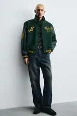 Cazadora estilo varsity con cuerpo de mezcla de lana en color verde oscuro y mangas de efecto piel a tono. Presenta cuello solapa, cierre frontal con botones a presión, bolsillos laterales de vivo y puños y bajo acanalados con rayas amarillas. Incluye bordados y parches decorativos en el frente con la inscripción Boston Archive.