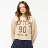 Buzo de cuello redondo en felpa color beige, con diseño estampado frontal con la inscripción 'NEW YORK 90 EAST VILLAGE' en color marrón. Presenta corte holgado y ruedo con terminación al corte.