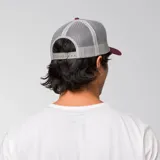 Gorra tipo trucker color bordó con panel frontal de sarga y paneles traseros de malla gris. Presenta el logo de la marca bordado en relieve en la parte frontal y visera curva.