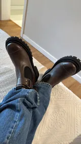 Bota tipo Chelsea de cuero marrón con suela track negra de goma. Presenta paneles elásticos laterales y una costura reforzada en la puntera. Incluye una correa de cuero desmontable con hebilla metálica.
