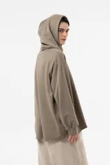 Buzo de punto oversize en color beige, con capucha, cordones largos ajustables y mangas largas amplias con puños elásticos. Presenta diseño asimétrico, más largo en la parte posterior.