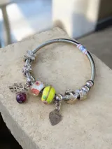 Pulsera rígida tipo brazalete con estructura de resorte, decorada con múltiples charms de metal plateado, cuentas de vidrio con motivos florales y geométricos, y dijes colgantes incluyendo un corazón con inscripción y un dije de árbol de la vida.