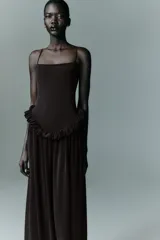 Vestido largo de color negro con escote recto y tirantes finos ajustables. Presenta un diseño con detalle de volados en la cintura que genera un efecto peplum y una falda con caída fluida.