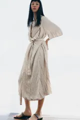 Vestido midi de Zara Woman Collection color beige, con cuello solapa, mangas por debajo del codo y diseño cruzado. Confeccionado en tejido plisado con brillo satinado, incluye un cinturón ajustable del mismo género con lazada en la cintura.