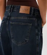 Pantalón de jean estilo baggy balloon, de tiro medio y corte amplio con diseño redondeado en las piernas. Presenta recortes laterales, cierre frontal con botón y cremallera, y bolsillos funcionales.