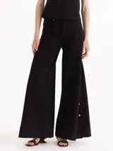 Pantalón de denim negro de tiro alto, confeccionado en algodón pima. Presenta silueta de pernera ancha con costuras frontales verticales y broches metálicos decorativos en los laterales de la botamanga. Su diseño incluye cierre frontal con botón metálico y cremallera oculta, además de bolsillos clásicos.