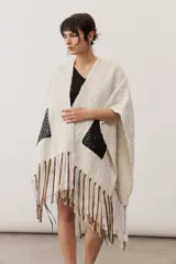 Ruana de punto color beige con detalles geométricos negros y flecos largos tricolores en el borde inferior.