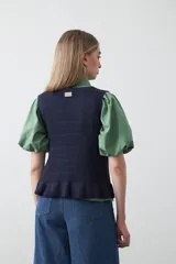 Chaleco tejido color verde musgo, con trama texturada, cuello a la base y cierre frontal con lazos. Presenta un ruedo con volado.