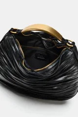 Bolso de mano negro de diseño fruncido con detalle de tiras y bolsillo interior con cremallera. Cuenta con un asa rígida metálica dorada y cierre principal mediante cremallera.