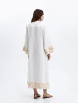 Vestido largo de lino blanco con cuello en V, mangas largas y bolsillos laterales. Presenta detalles de bordado geométrico en tono dorado en los puños y en el bajo.