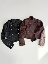 Campera de estilo militar en color marrón oscuro, confeccionada en gamuza. Presenta un diseño cruzado con doble fila de botones metálicos grabados, cuello alto y charreteras en los hombros.