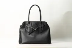 Cartera shopper negra de cuero sintético con asas largas para el hombro, detalles de correas frontales con hebilla y marca She Mona grabada en el frente.