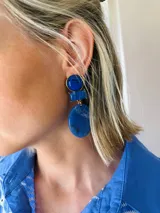 Caravanas largas compuestas por tres piezas de acrílico en tono azul: un círculo superior, un rectángulo central y un óvalo inferior, unidos por herrajes dorados.