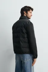 Cazadora acolchada de cuello subido, color negro, con mangas largas de tejido elástico. Incluye bolsillos en la cadera, un bolsillo interior y bajo con elástico. Cierre frontal de cremallera.