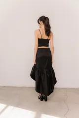 Pollera midi de tafeta color negro, con diseño asimétrico de corte mullet (más corta adelante y larga atrás), tiro alto y detalle de volado fruncido en el ruedo.