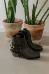 Botinetas de cuero negro con diseño estilo western, punta cuadrada y taco bajo con acabado desgastado. Presentan una tira decorativa con estribo metálico desmontable alrededor del tobillo.