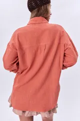 Camisa color coral de corte holgado, con cuello clásico, cierre frontal con botones, mangas largas con puños abotonados, dos bolsillos de parche en el pecho y ruedo asimétrico.