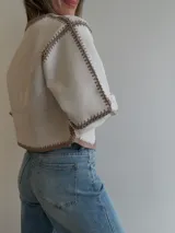 Chaqueta de efecto gamuza color tostado, con diseño abierto sin cierres y detalles de costuras artesanales tipo crochet en color blanco en los bordes, el frente, el cuello y las mangas.