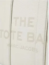 Cartera tipo tote bag Marc Jacobs modelo The Leather Small Tote Bag, color negro, confeccionada en cuero graneado con el texto 'THE TOTE BAG' grabado en relieve al frente. Cuenta con asas superiores, correa ajustable y desmontable, y herrajes metálicos dorados.