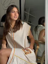Vestido corto beige de algodón con cuello camisero y cierre frontal de botones. Presenta mangas cortas y detalles bordados a contratono en la zona del pecho y en los bolsillos, además de un volado en el ruedo.
