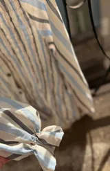 Camisa de manga larga con diseño a rayas verticales en tonos azul, gris y crema. Presenta un cuerpo con frunces elásticos decorativos, cuello con volados y puños con elástico y terminación en volado. Incluye cierre frontal con botones.