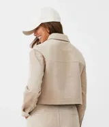 Campera corta color beige de pana con cuello camisero, cierre frontal con botones, bolsillos de parche en el pecho y mangas largas con puños abotonados.