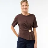 Camiseta marrón de manga corta con cuello redondo, diseño drapeado al frente y un botón metálico decorativo en la parte inferior.