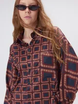 Camisa de seda con estampado geométrico de cuadrados en tonos marrones y azul oscuro. Presenta cuello camisero, cierre frontal con botones, bolsillo en el pecho y mangas largas con caída ligera.