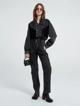 Mono de denim negro con cierre frontal de botones, cintura elástica y detalle de flecos en la parte superior delantera. Presenta mangas largas con puños elásticos y corte recto en las piernas.