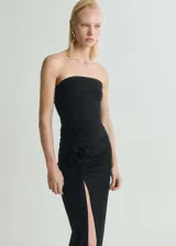 Vestido largo negro de diseño entallado, con escote palabra de honor y sin mangas. Presenta un detalle de flores 3D en la cintura y una apertura lateral en el bajo.
