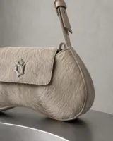 Cartera de mano de cuero vacuno con pelo largo en tono crudo y vainilla, con forma curva y solapa frontal con cierre magnético y aplique metálico con monograma. Cuenta con una correa ajustable corta para llevar al hombro o en la mano.