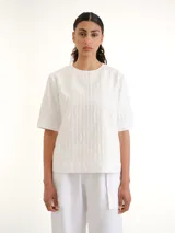 Blusa blanca de manga corta y cuello redondo, con diseño de rayas verticales texturizadas y detalles sutiles de flecos.