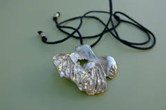 Collar con dije de metal plateado con forma irregular y textura rugosa, con un orificio en el centro. El dije cuelga de un cordón negro ajustable.