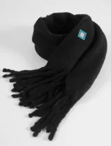 Bufanda de textura suave y acabado afelpado, color negro, con flecos en los extremos y un pequeño parche cuadrado decorativo con logo en color verde y azul.