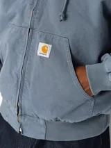 Campera OG Active de Carhartt WIP, color azul grisáceo, con capucha integrada, cierre frontal con cremallera metálica, bolsillos tipo canguro y parche con el logo de la marca en el bolsillo inferior.