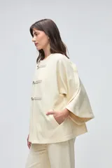 Capa de lana fina sin teñir, con diseño de corte amplio y mangas tipo kimono. Presenta cierre frontal mediante tres alamares de cuero en tono marrón.