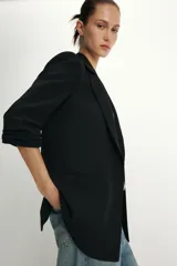 Blazer negro con cuello solapa con muescas, hombreras y forro interior a tono. Presenta manga por debajo del codo con detalle de drapeado y falsos bolsillos de vivo en el delantero, con cierre frontal de un solo botón dorado.