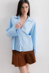 Camisa azul claro de algodón, con cuello camisero, mangas largas y puños anchos. Cierre frontal con botones y un bolsillo de parche en el lateral.