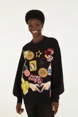 Buzo de punto negro, de corte holgado, con cuello redondo y mangas largas con puños ajustados. Presenta múltiples apliques bordados con motivos tropicales y coloridos en la parte frontal.