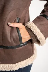 Campera tipo gamulán de corte oversize, color marrón con interior y cuello de corderito sintético en tono crema. Presenta detalles de ribetes en cuero sintético negro a lo largo de la prenda, cierre frontal metálico y puños tejidos.