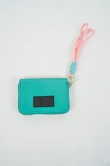 Billetera pequeña color turquesa con cierre perimetral y parche frontal negro con logo. Incluye una correa de muñeca de cordón rosado con detalle de hilo celeste.