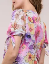 Blusa de gasa liviana con estampado floral en tonos violeta y rosado, cuello redondo y mangas cortas con detalle de lazo en los puños.
