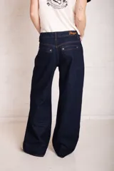 Pantalón de jean azul oscuro, de corte holgado y tiro bajo, con pierna ancha y efecto lavado. Presenta costuras en color ocre y parche con logo vegano en la parte trasera. Confeccionado en denim rígido sin elasticidad.