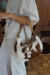 Bolso shoulder bag de cuero con pelo con estampado de vaca en tonos marrón y blanco. Cuenta con doble asa: una correa de cuero con tachas y una cadena metálica desmontable.