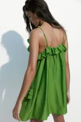 Vestido evasé corto de color verde, confeccionado en mezcla de viscosa con leve brillo. Presenta breteles finos ajustables, escote en U con cordón de ajuste y volados decorativos fruncidos en la parte superior.