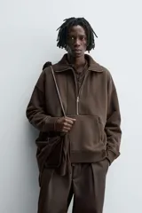 Sudadera de corte relajado color marrón oscuro, con cuello subido y cierre frontal de cremallera. Presenta manga larga, bolsillo frontal tipo canguro y acabados en rib en puños y ruedo.