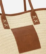 Bolso de playa tipo tote bag, tejido en fibra de papel color natural, con bordes y detalles en cuero sintético marrón. Posee asas de hombro en cuero sintético marrón y un parche cuadrado con logo grabado en el frente.