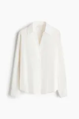 Blusa blanca de tela con brillo, cuello clásico, escote en V con botones frontales, mangas largas con puños sencillos y bajo redondeado.